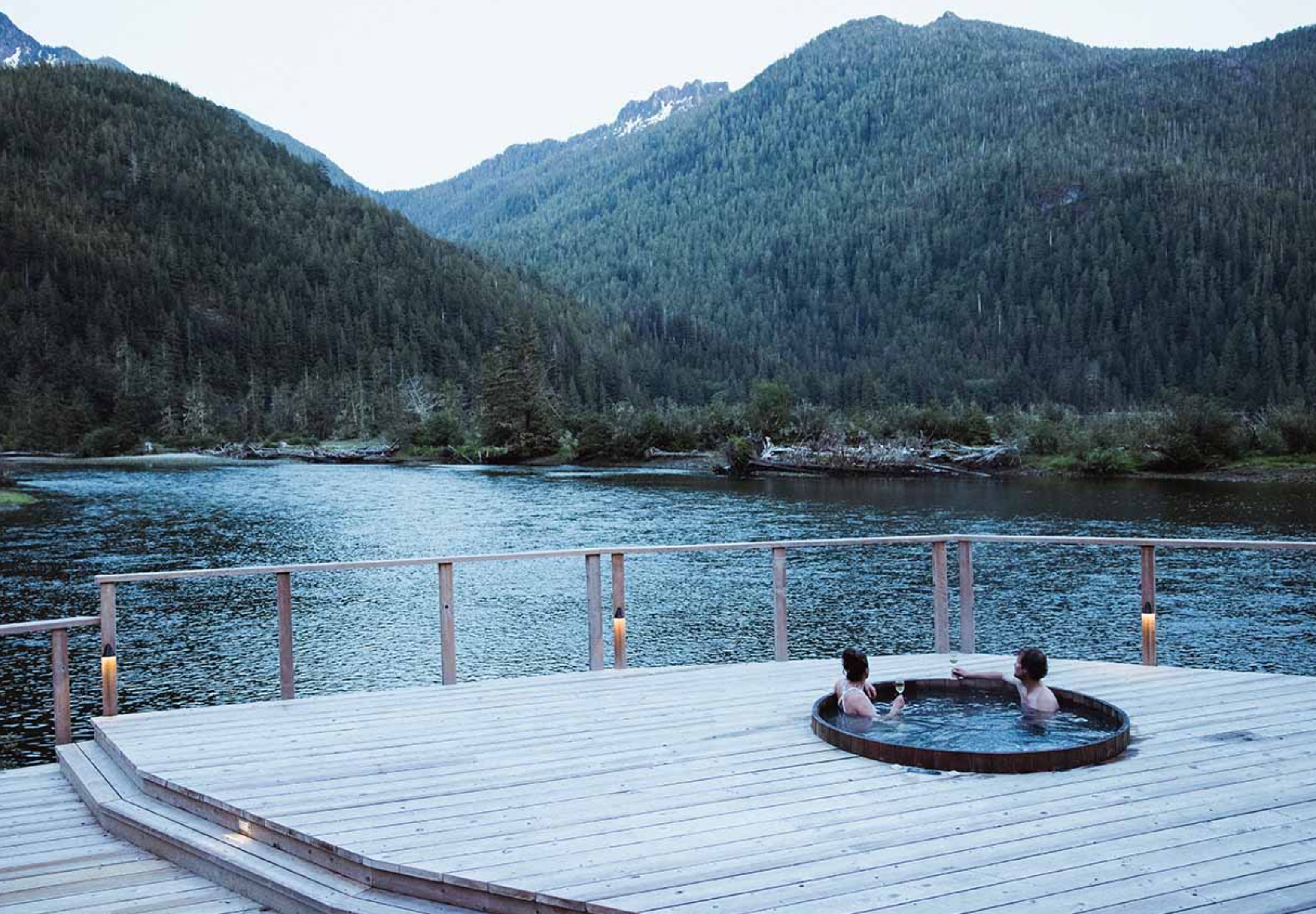 Two people in hot tub on wooden deck looking over lake and mountain view.