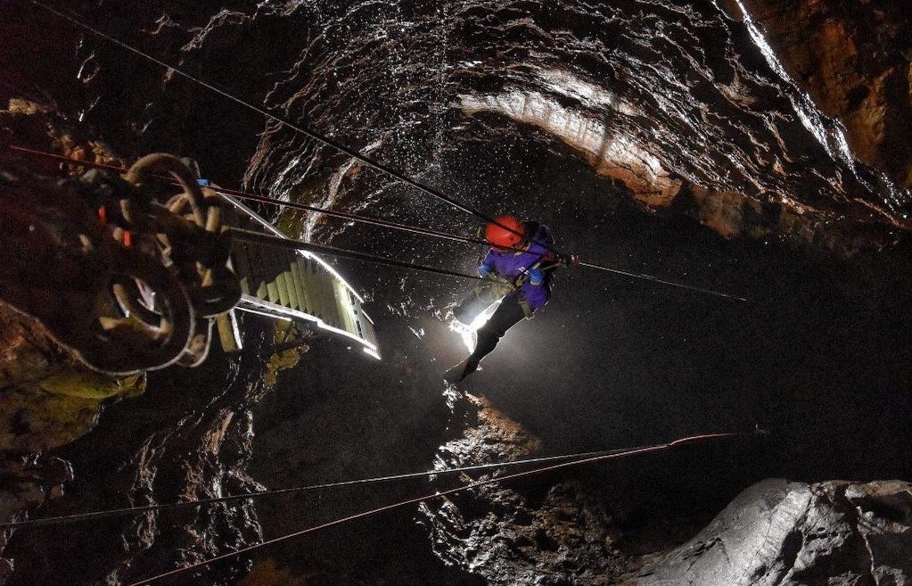 Person diving into a deep cave attached to wired lowering device.